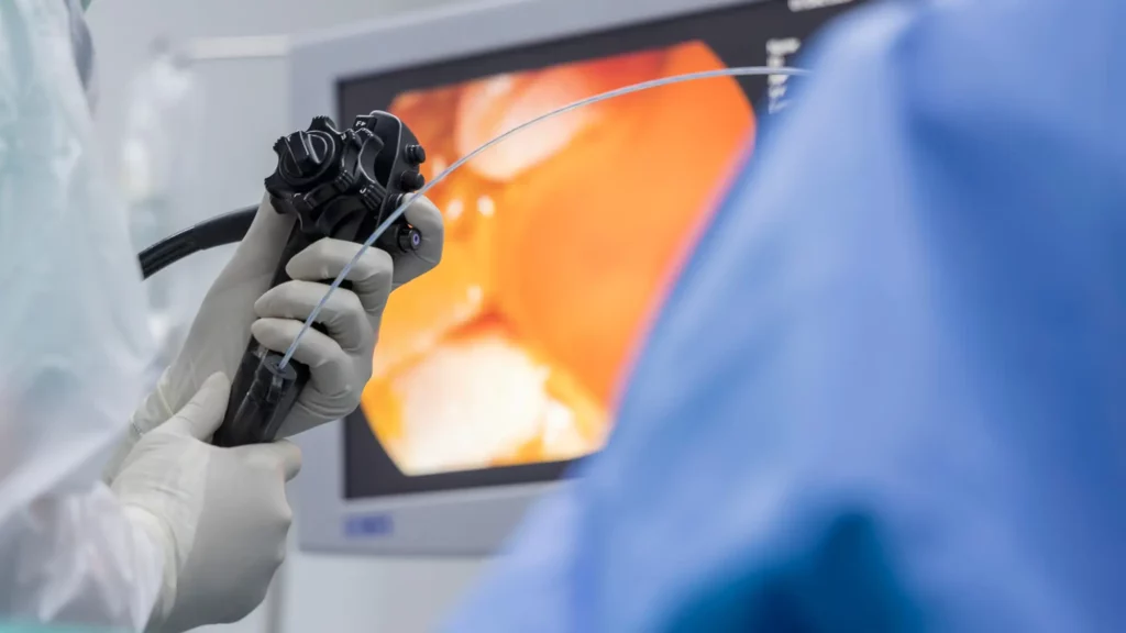 Medical professional holding an endoscope performing a procedure. The monitor in the background displays a magnified internal view depicting orange-hued tissues.