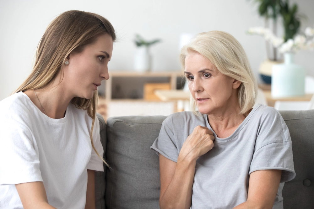 Two women sit closely on a couch indoors, engaged in a serious conversation. The younger woman with long brown hair listens intently while the older woman with short blonde hair gestures as she speaks. The background is softly blurred.