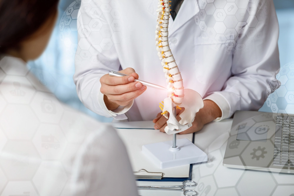 A doctor in a white lab coat uses a pen to point at a model of a human spine on a desk. A clipboard and laptop are nearby. Another person listens in the foreground. Hexagonal patterns overlay the image, suggesting medical themes.
