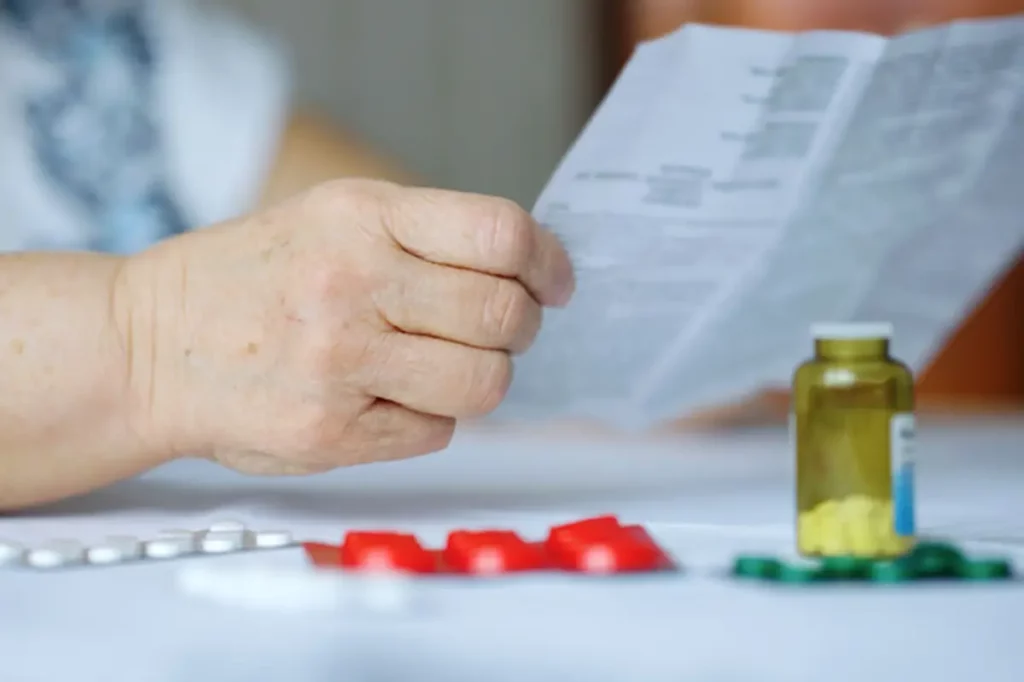 Close-up of an elderly persons hand holding a paper reading medication instructions. Beside them is a table with various blister packs of pills and a small transparent bottle with yellow tablets.