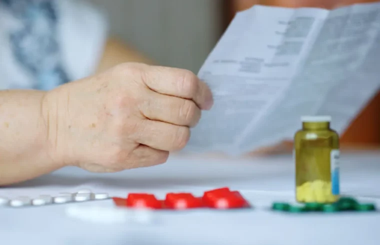 Close-up of an elderly persons hand holding a paper reading medication instructions. Beside them is a table with various blister packs of pills and a small transparent bottle with yellow tablets.