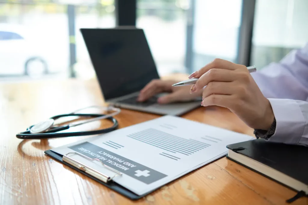 A person wearing a white coat is working on a laptop at a wooden desk. A stethoscope, a clipboard with medical documents, and a notebook are also on the desk. The scene is well-lit with a window in the background.