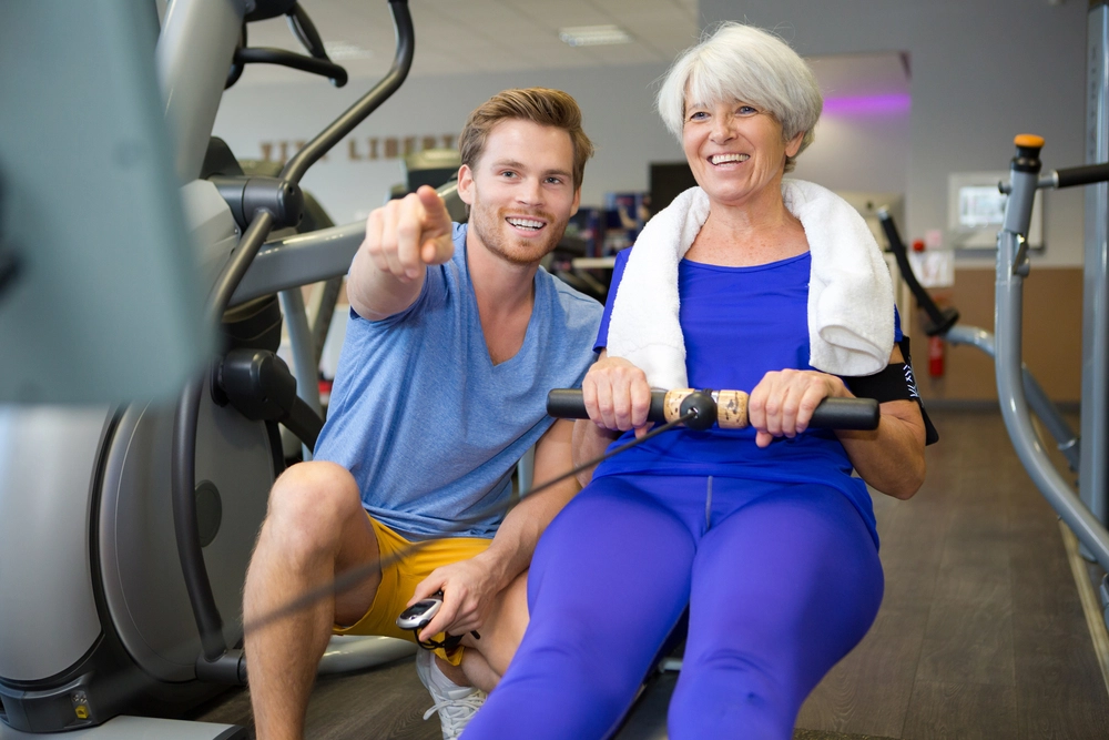 A smiling woman in workout clothes uses a rowing machine with a man beside her pointing forward in a gym setting.